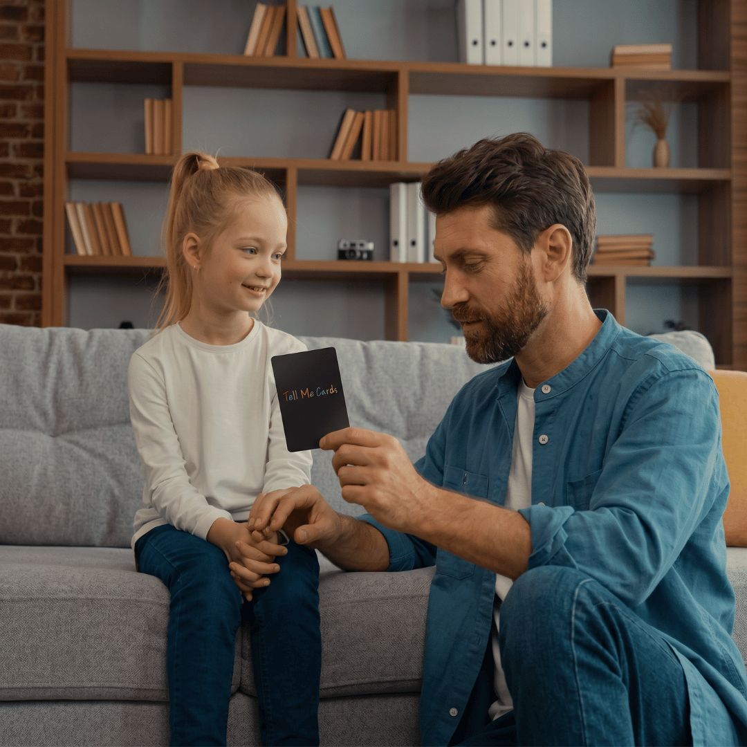 Father reading a Tell Me Cards conversation card to his young daughter on the couch at home.