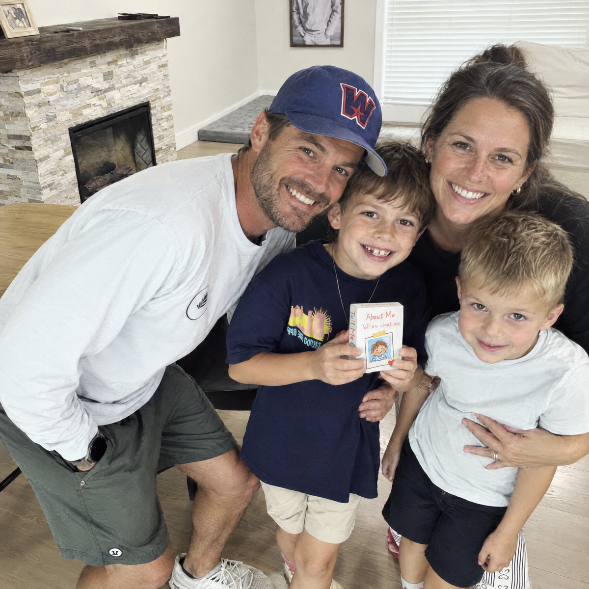A family of four smiling together at home, the older boy holding the Tell Me Cards About Me deck