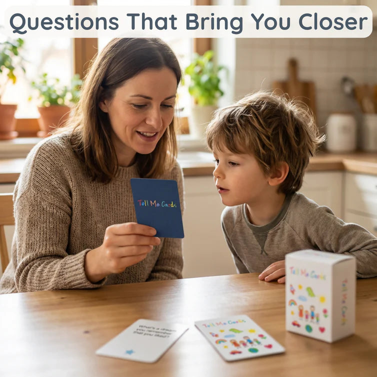 A mother holding up a Tell Me Cards question card to her young son at a kitchen table, sharing a conversation